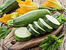 Eine Zucchini liegt auf einem Holzbrett. Daneben liegen mehrere Zucchini-Scheiben und Zucchini-Blüten.