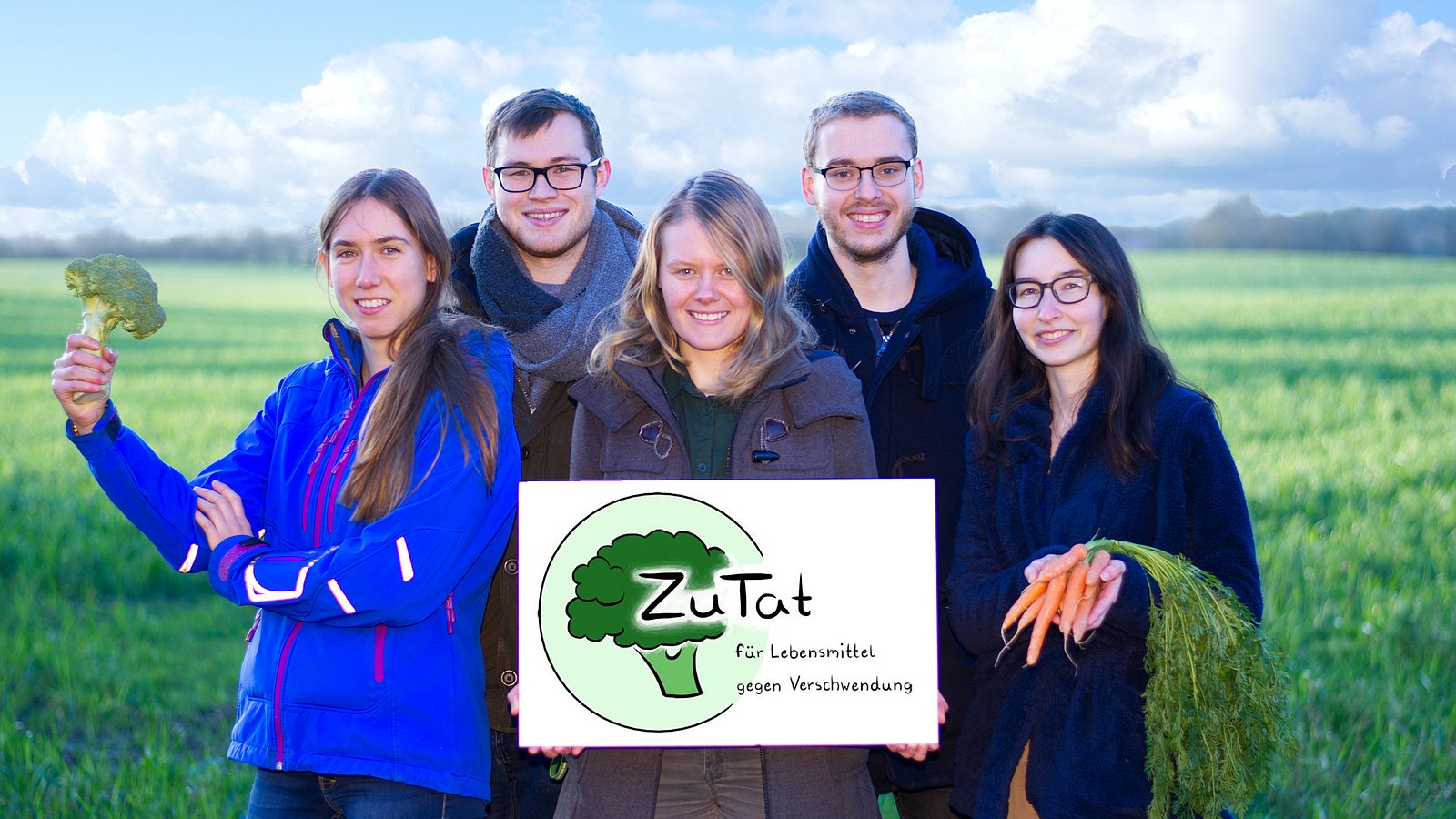 Das Team von ZuTat steht auf einem Feld und hält Gemüse in der Hand. Es hält ein Schild mit dem Logo in die Kamera.