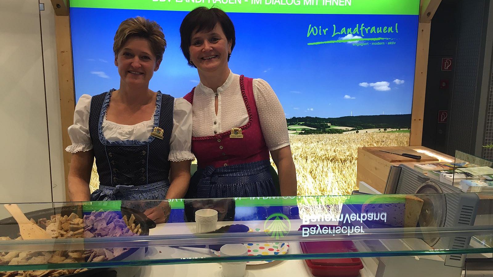 Zwei Frauen in Trachtenkleidern stehen an einem Essensstand des Bayerischen Bauernverbandes.