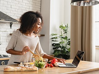Eine Frau kocht und schaut dabei auf ihren Laptop.