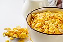 Eine Schüssel Cornflakes, neben der ein Löffel mit trockenen Cornflakes liegt. Daneben steht ein Glaskrug mit Milch.