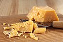 Ein Stück Hartkäse liegt auf einem Holzbrett neben einem Käsemesser.
