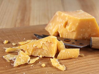 Ein Stück Hartkäse liegt auf einem Holzbrett neben einem Käsemesser.