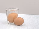 Ein braunes Ei liegt neben einem Glas, in dem ein weiteres braunes Ei in Wasser liegt.
