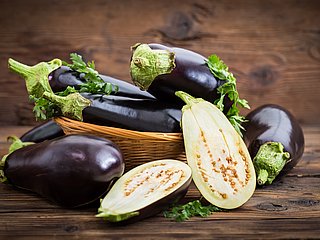 Vier Auberginen liegen drapiert in und um einen Korb herum. Zwei Auberginen-Hälften liegen davor 