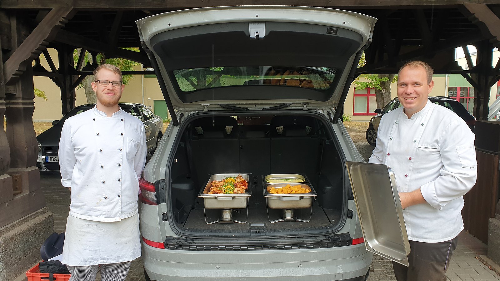 Zwei Menschen vor einem Auto-Kofferraum, indem zubereitete Speisen in einer Schüssel liegen