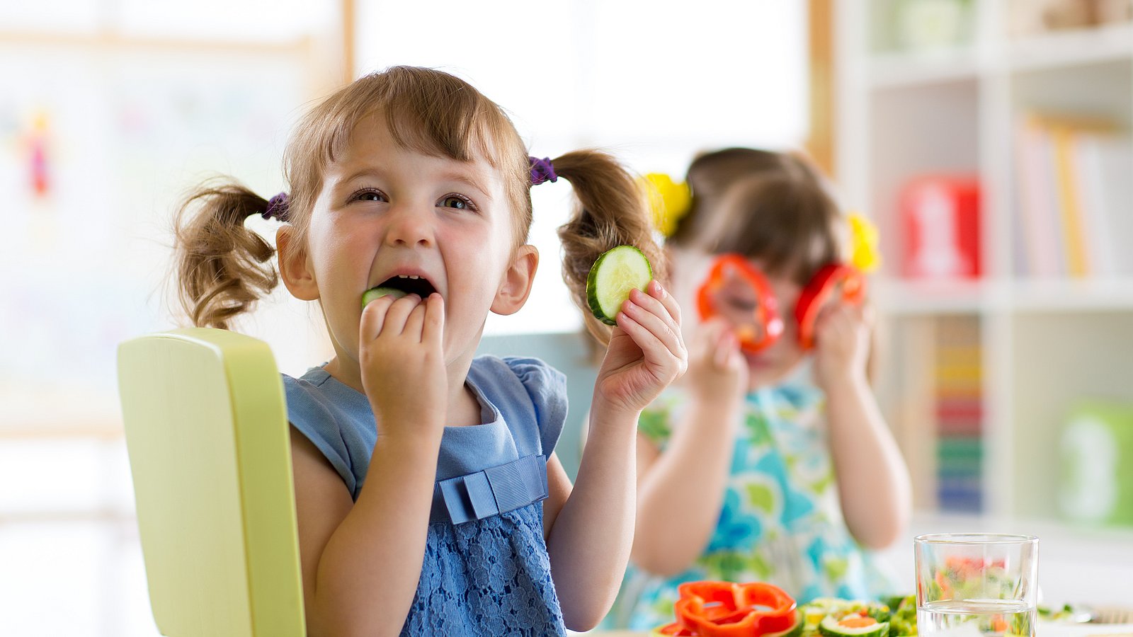 Zwei kleine Mädchen sitzen am Tisch und spielen mit Gurkenscheiben und Paprikaringen.