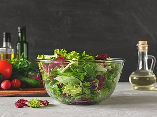 Salat wird in einer großen Glasschüssel präsentiert. Daneben stehen mehrere Flaschen mit Öl und Essig und weiteres Gemüse liegt auf einem Holzbrett.