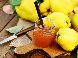 Gelbe Quitten liegen auf einem Holzbrett. Davor steht ein Glas mit Quitten-Marmelade und liegt ein Messer.