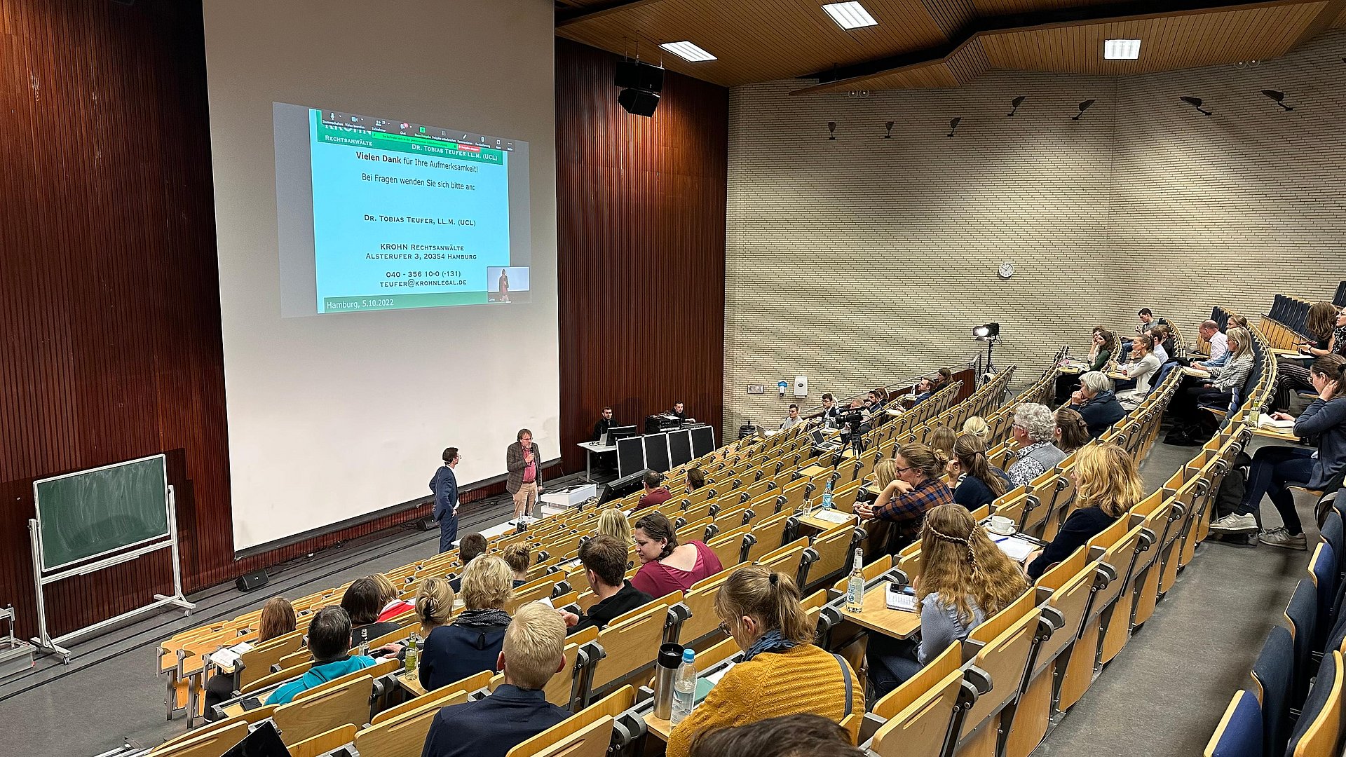 Das Bild zeigt einen halbvollen Hörsaal von oben. Vorne steht ein älterer Herr und hält einen Vortrag