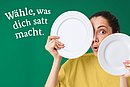 Schrift: Wähle, was dich satt macht. Frau hält zwei unterschieldich große weiße Teller vor das Gesicht