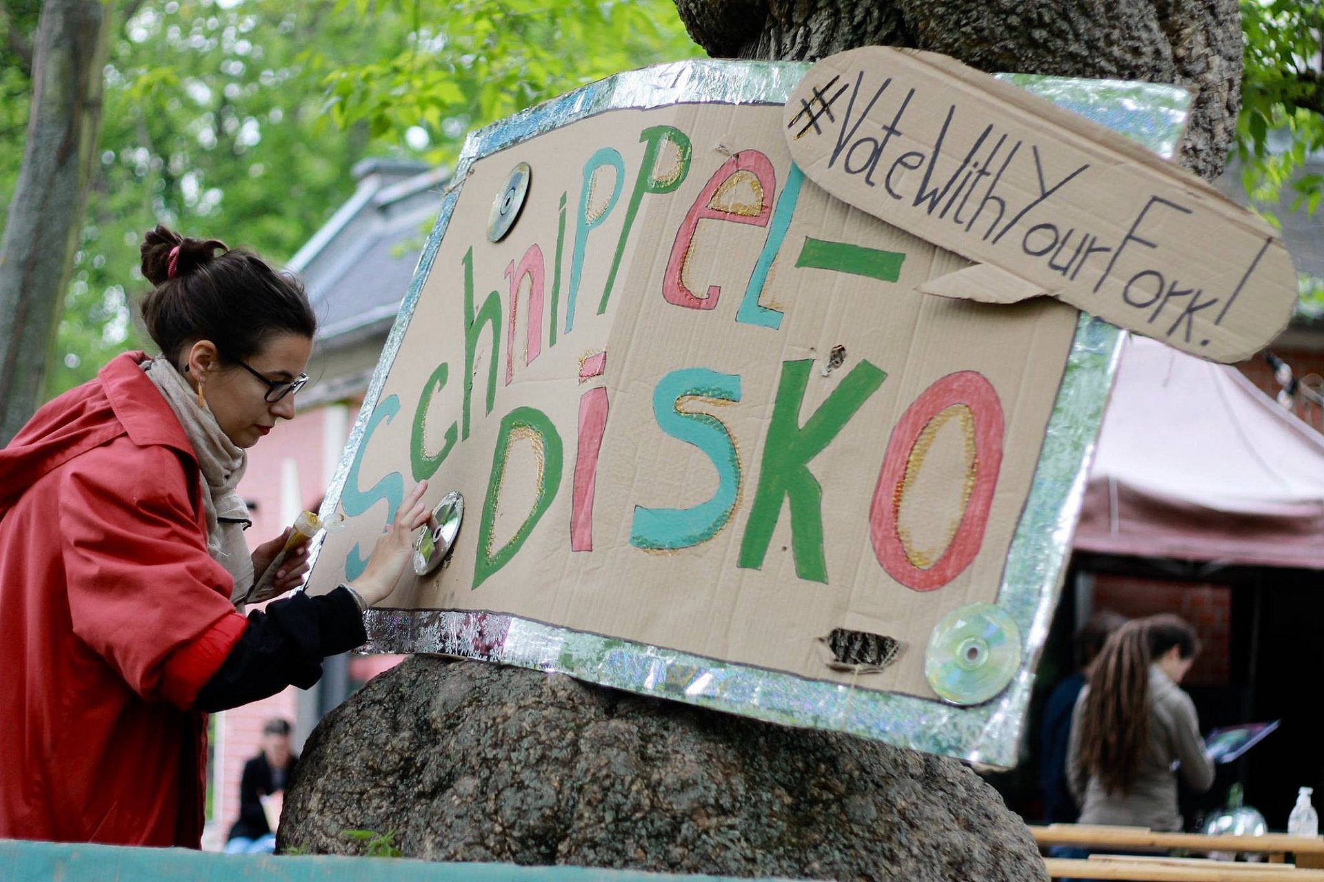 Das Foto zeigt eine Frau, die ein selbst gemachtes Plakat bearbeitet, das an einem Baum angebracht ist. Auf dem Plakat steht: Schnippel-Disko.