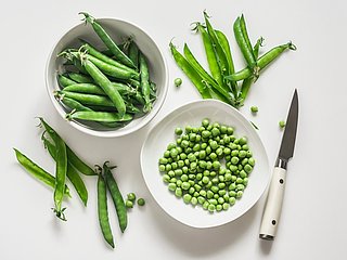 Eine Schüssel mit Erbsen und eine Schüssel mit Erbsenschoten. Daneben liegt ein Messer und weitere Erbsenschoten.