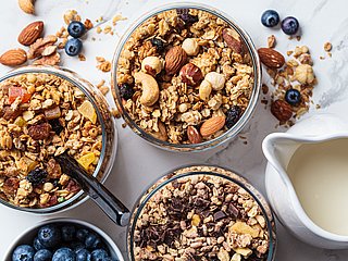 Verschiedene Müslisorten werden in drei Schüsseln präsentiert. Daneben steht eine Schüssel mit Brombeeren und ein Krug mit Milch.