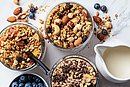 Verschiedene Müslisorten werden in drei Schüsseln präsentiert. Daneben steht eine Schüssel mit Brombeeren und ein Krug mit Milch.