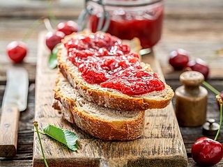 Zwei Brotscheiben bestrichen mit Marmelade liegen auf einem Holzbrett. Dahinter steht ein Einweck-Glas mit Marmelade und daneben liegt ein Messer.