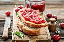 Zwei Brotscheiben bestrichen mit Marmelade liegen auf einem Holzbrett. Dahinter steht ein Einweck-Glas mit Marmelade und daneben liegt ein Messer.
