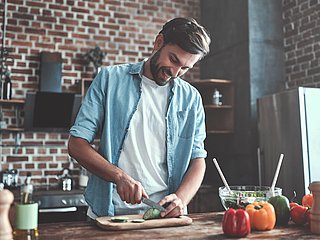 Ein junger Mann im blauen Hemd steht in einer Küche und schneidet Gemüse