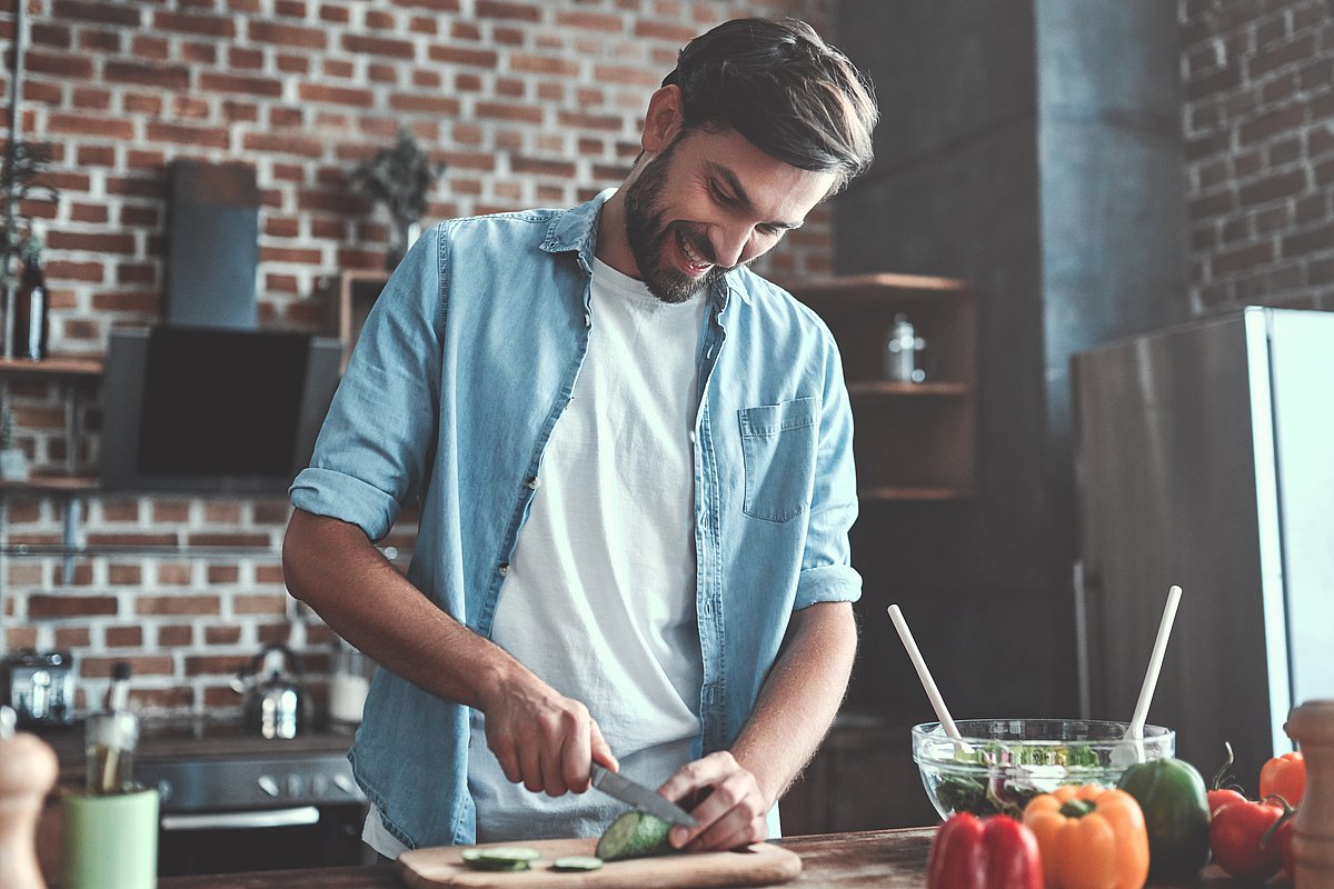 Ein junger Mann im blauen Hemd steht in einer Küche und schneidet Gemüse