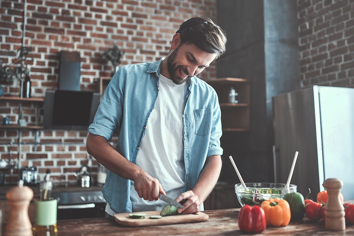 Ein junger Mann im blauen Hemd steht in einer Küche und schneidet Gemüse