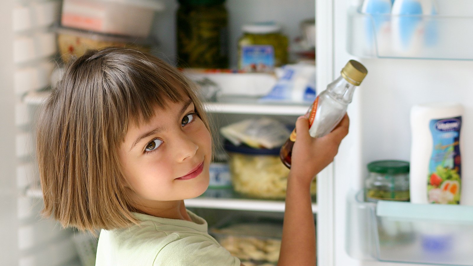 Ein Kind hält eine Flasche in der Hand und steht vor einem offenen Kühlschrank. 