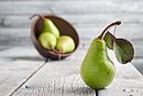 Eine grüne Birne mit Blatt im Vordergrund. Im Hintergrund drei grüne Birnen in einem Korb.