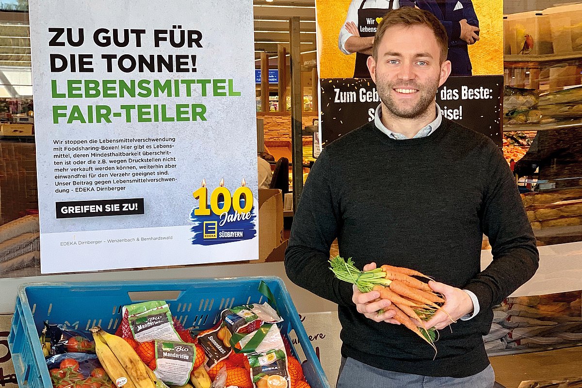 Ein Mann steht mit einem Bündel Möhren in der Hand haltend neben einer Kiste mit abgelaufenen aber, noch verwendbaren Lebensmitteln im Supermarkt. 