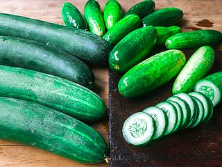 Mehrere Gurken liegen auf einem Holzbrett. Daneben liegt eine in Scheiben geschnittene Gurke.