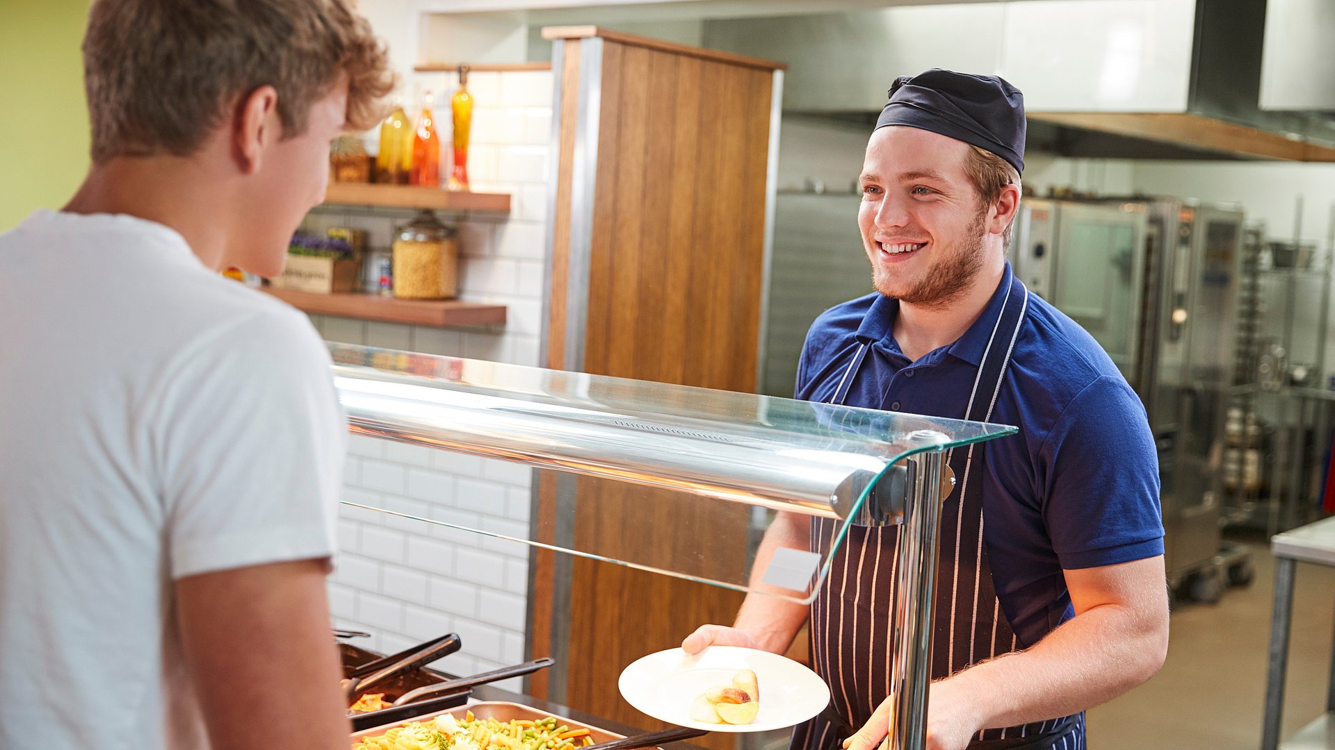 Das Bild zeigt einen zwei Männer in einer Kantine. Ein Mann holt was zu essen an der Theke, der andere bedinet ihn mit einem lächeln im Gesicht.