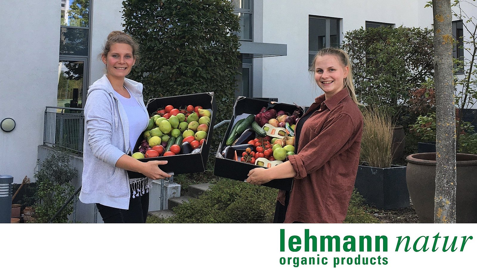 Zwei Mitarbeiterinnen von lehmann natur stehen vor einem Haus und halten Obstkisten in die Kamera.