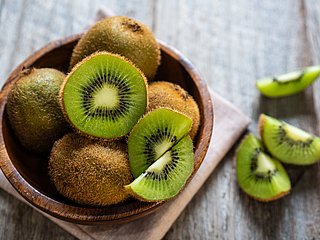 Einige Kiwis liegen, teilweise geschnitten, teilweise im Ganzen, in einer Holzschale.