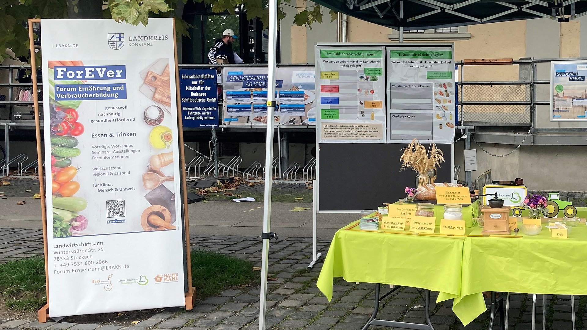 Foto eines Standes auf dem Biomarkt mit einem Aufsteller mit Informationen und einem Tisch, auf dem unterschiedliche Lebensmittel und Gegenstände stehen.