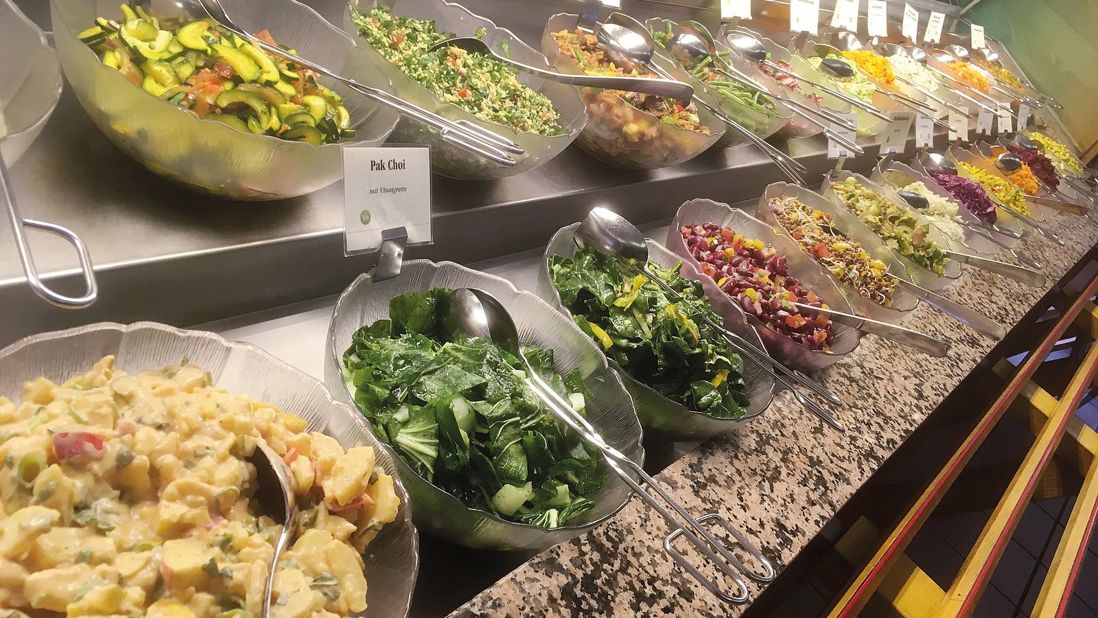 Auf einer Salattheke stehen Schüsseln mit verschiedenen Salatvariationen. 