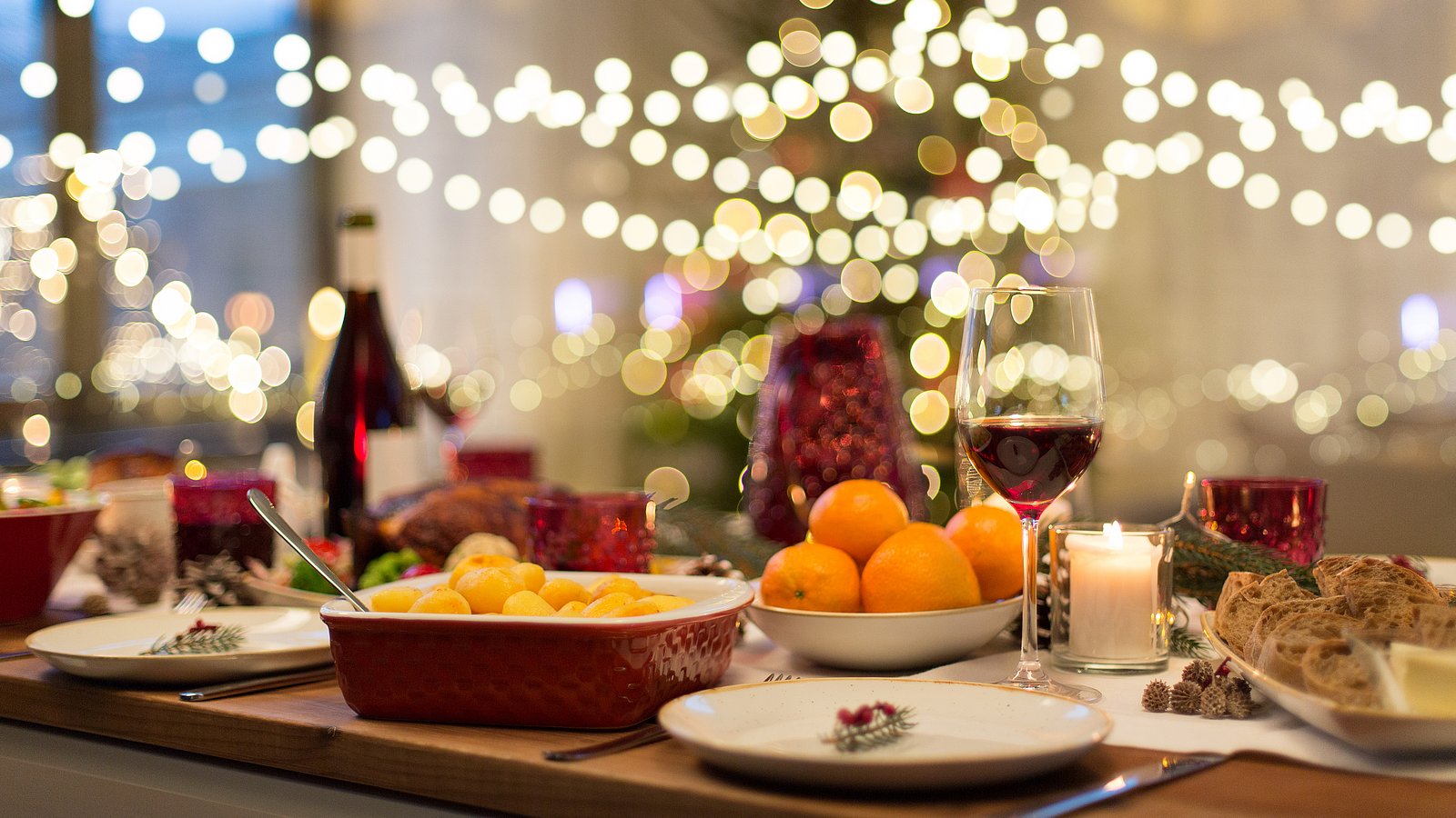 Festlich gedeckte Weihnachtstafel mit Orangen, Kartoffeln und Brot. Im Hintergrund hängen Lichterketten.