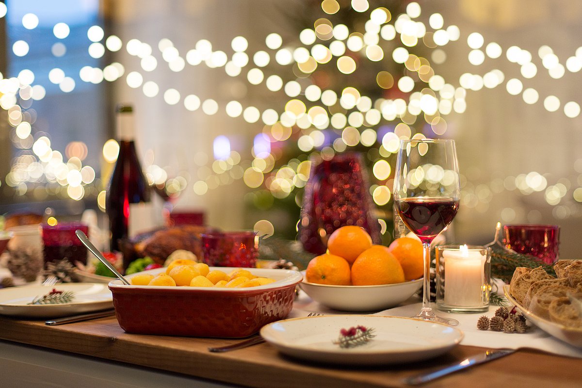Festlich gedeckte Weihnachtstafel mit Orangen, Kartoffeln und Brot. Im Hintergrund hängen Lichterketten.