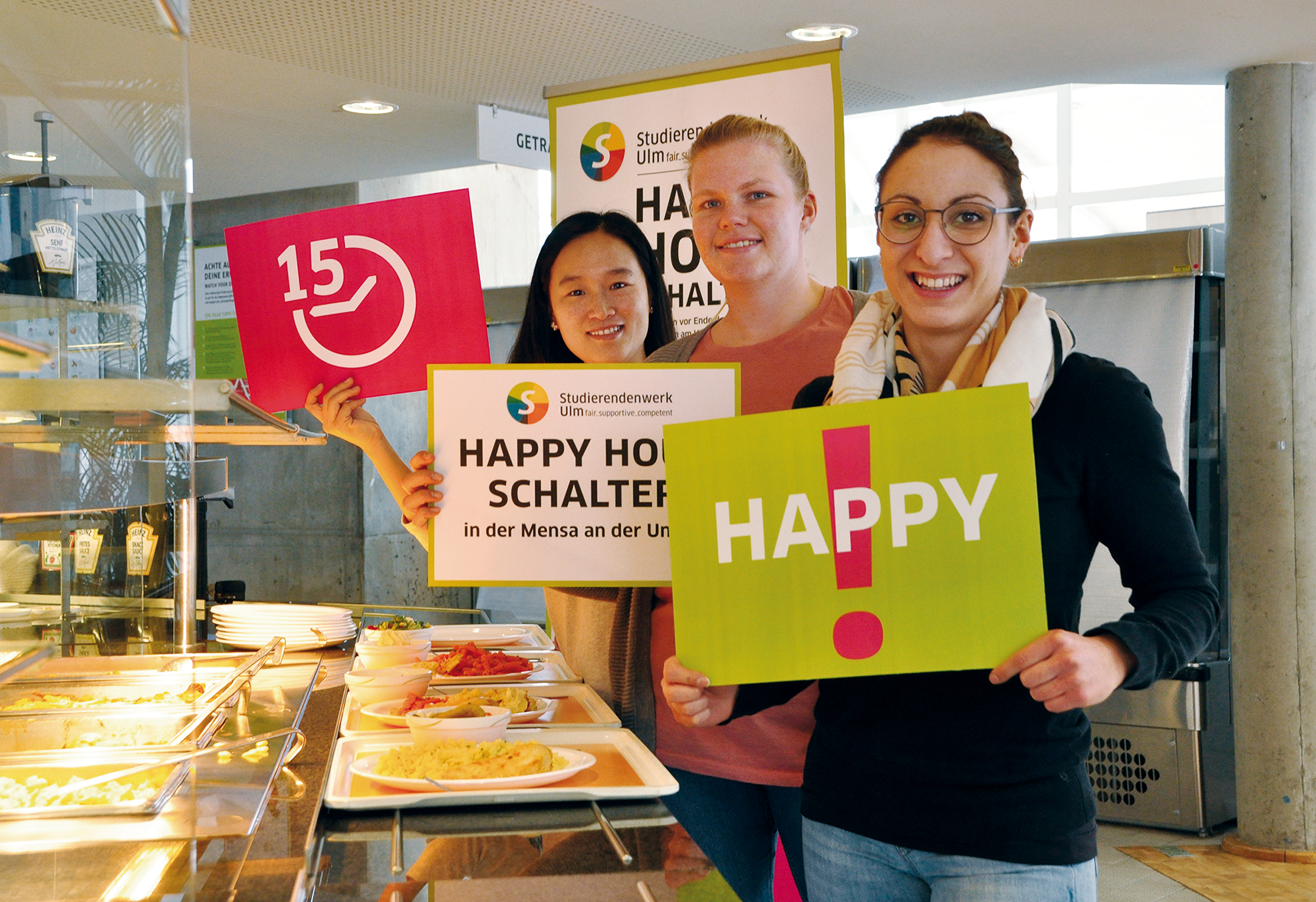 Drei Personen halten unterschiedliche Plakate in der Hand in einer Mensa. Auf einem Plakat steht "Happy Hour". 